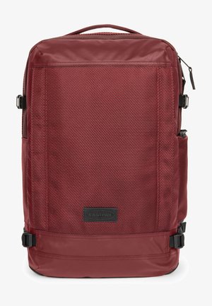 Red fabric backpack with a textured surface, flat top, side pocket, adjustable straps, and a brand logo patch on the front.