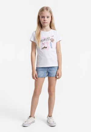 Ragazza bionda in piedi che indossa una t-shirt bianca con grafica, pantaloncini di jeans e sneakers bianchi con accenti maculati su sfondo bianco.