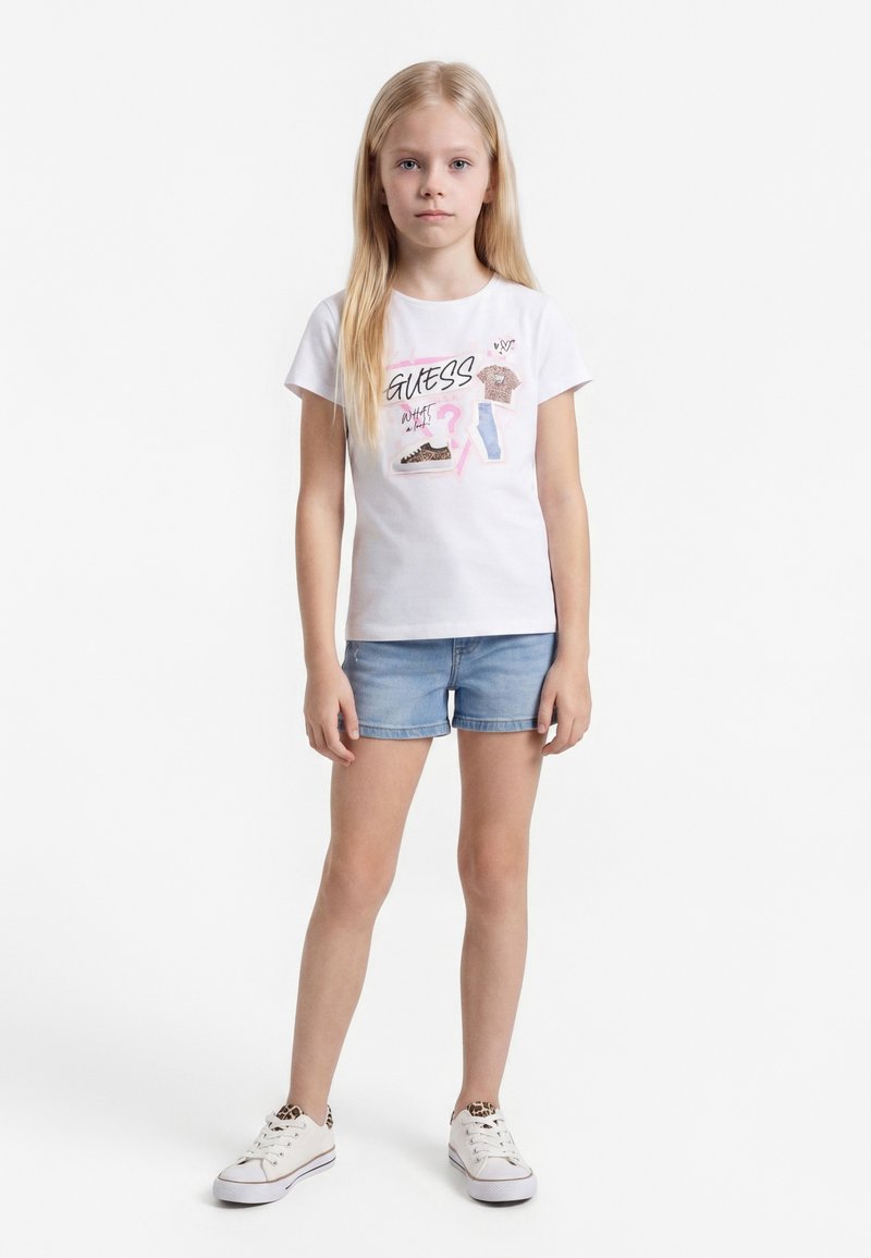 Blond meisje staat, draagt een wit T-shirt met grafische print, denimshorts en witte sneakers met luipaardprint accenten tegen een witte achtergrond.