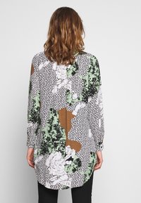 Langærmet skjorte med en blanding af blomster- og geometriske mønstre i grøn, hvid, sort og brun. Har en buet nederkant og knaplukning ved ærmerne.