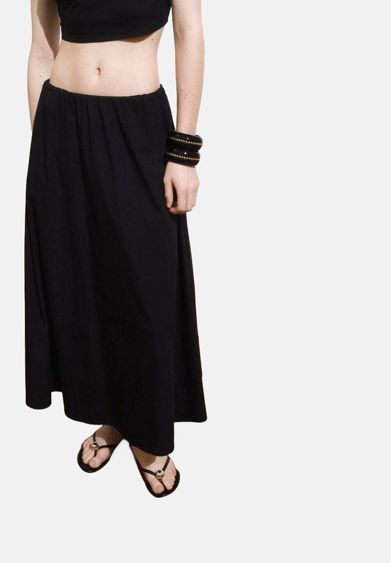 Jupe noire longueur cheville avec une taille élastique, en tissu lisse ; associée à des sandales plates à lanières et de gros bracelets noirs.