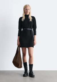 Schwarzes Strickkleid mit hohem Kragen und kurzen Ärmeln, tailliert mit einem dünnen Gürtel. Kombiniert mit schwarzen Schnürstiefeln und einer braunen Tragetasche.