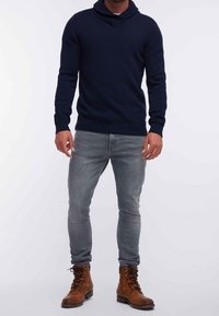 Pull-over bleu marine en maille avec col châle, jeans gris slim, et bottines lacées couleur fauve au fini cuir lisse.