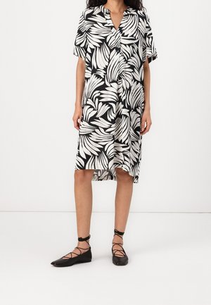 Femme portant une robe aux manches courtes, longueur genoux, à motif de feuilles noir et blanc, avec des ballerines noires lacées, sur un fond blanc uni.