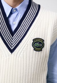 Biała dzianina kamizelki z granatowymi paskami wzdłuż dekoltu w serek, z zielonym naszywką z logo krokodyla i fakturowanym prążkowaniem. Pod spodem jasnoniebieska koszula.