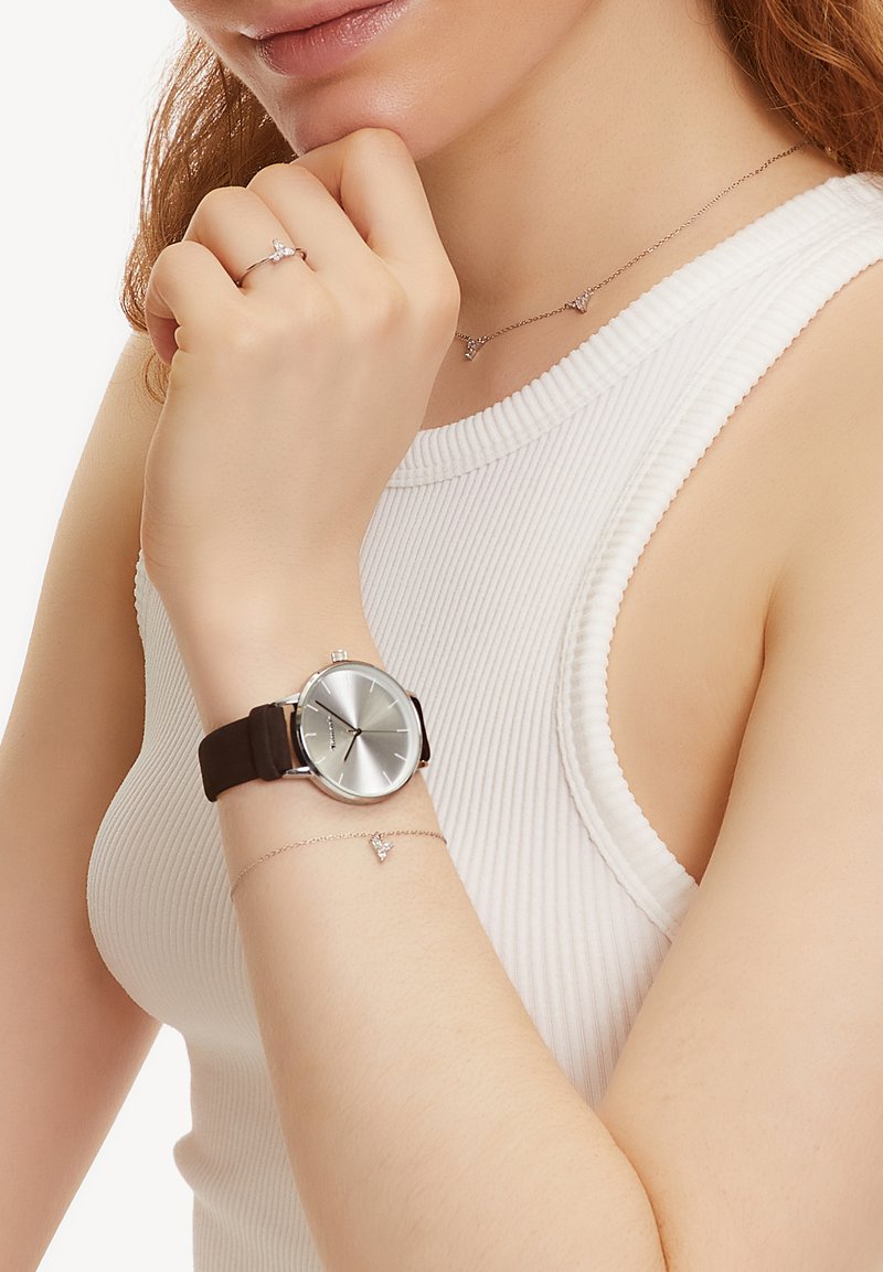 Silberne Uhr mit rundem Zifferblatt und braunem Silikonarmband, getragen mit einem filigranen Silberarmband, das kleine dreieckige Akzente aufweist. Weißes geripptes Oberteil.
