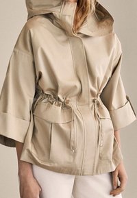 Beżowa, wodoodporna kurtka z kapturem, ściągaczem w talii, szerokimi rękawami oraz dwoma przednimi kieszeniami. Gładka tekstura z błyszczącym wykończeniem.