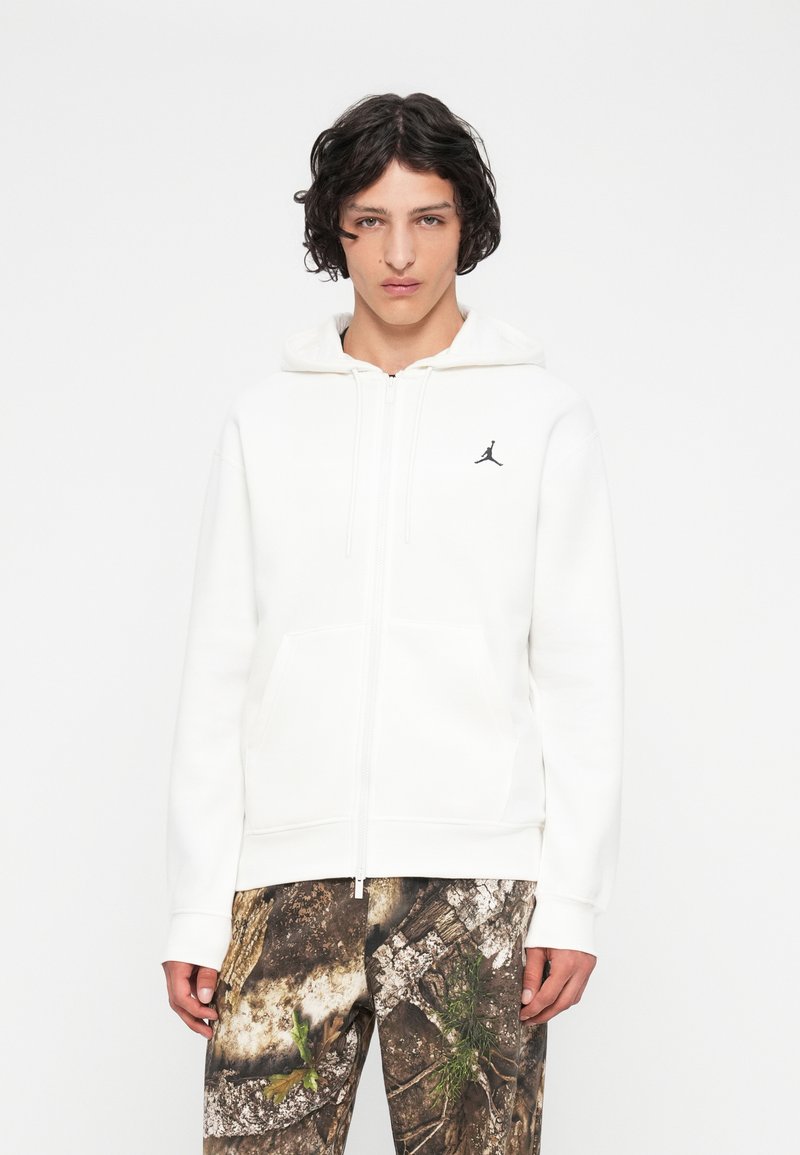 Giovane con capelli ricci e scuri che indossa una felpa bianca con zip e un piccolo logo nero, insieme a pantaloni con motivo mimetico.
