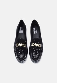 Μαύρα μοκασίνια από σουέτ με γυαλιστερή χρυσή αγκράφα σε σχήμα καρδιάς και διακοσμητικά με στρας διατεταγμένα σε μοτίβο βαθμίδωσης.