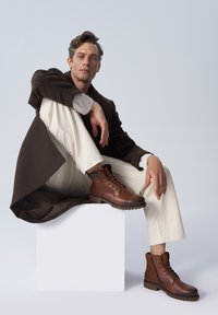 Brązowe skórzane botki za kostkę, podniesiona podeszwa, okrągły czubek, sznurowana konstrukcja z bocznym zamkiem błyskawicznym, teksturowane wykończenie, noszone z kremowymi spodniami i ciemnym płaszczem.