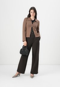 Cardigan marrone con zip, top in pizzo nero, pantaloni neri larghi, tacchi grigi e una piccola borsa nera di tessuto. Sfondo bianco semplice.