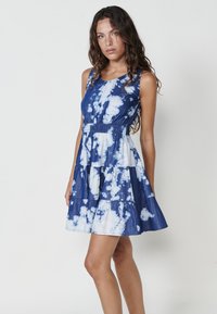 Plava haljina s tie-dye uzorkom, pripijenog gornjeg dijela, okruglog izreza i lepršavim rubom. Ima elastičnu struk i lagani materijal s različitim uzorcima.