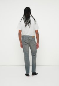 Bărbat cu păr lung împletit, purtând un tricou gri deschis, blugi gri decolorati și adidași negri, stând cu fața spre un perete alb.