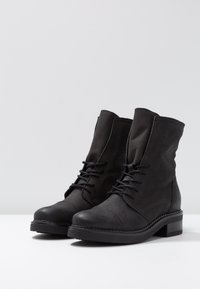Bottes noires à la cheville en cuir mat avec un bout arrondi, un laçage sur le devant et un petit talon épais. Présente des coutures le long de la semelle.