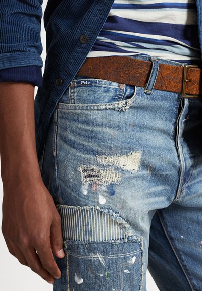 Tuvplāns cilvēka, kurš valkā saplēstas, nodilušas zilas džinsa bikses ar krāsas plankumiem, brūnu ādas jostu un zilu svītrainu kreklu zem zilas jakas.