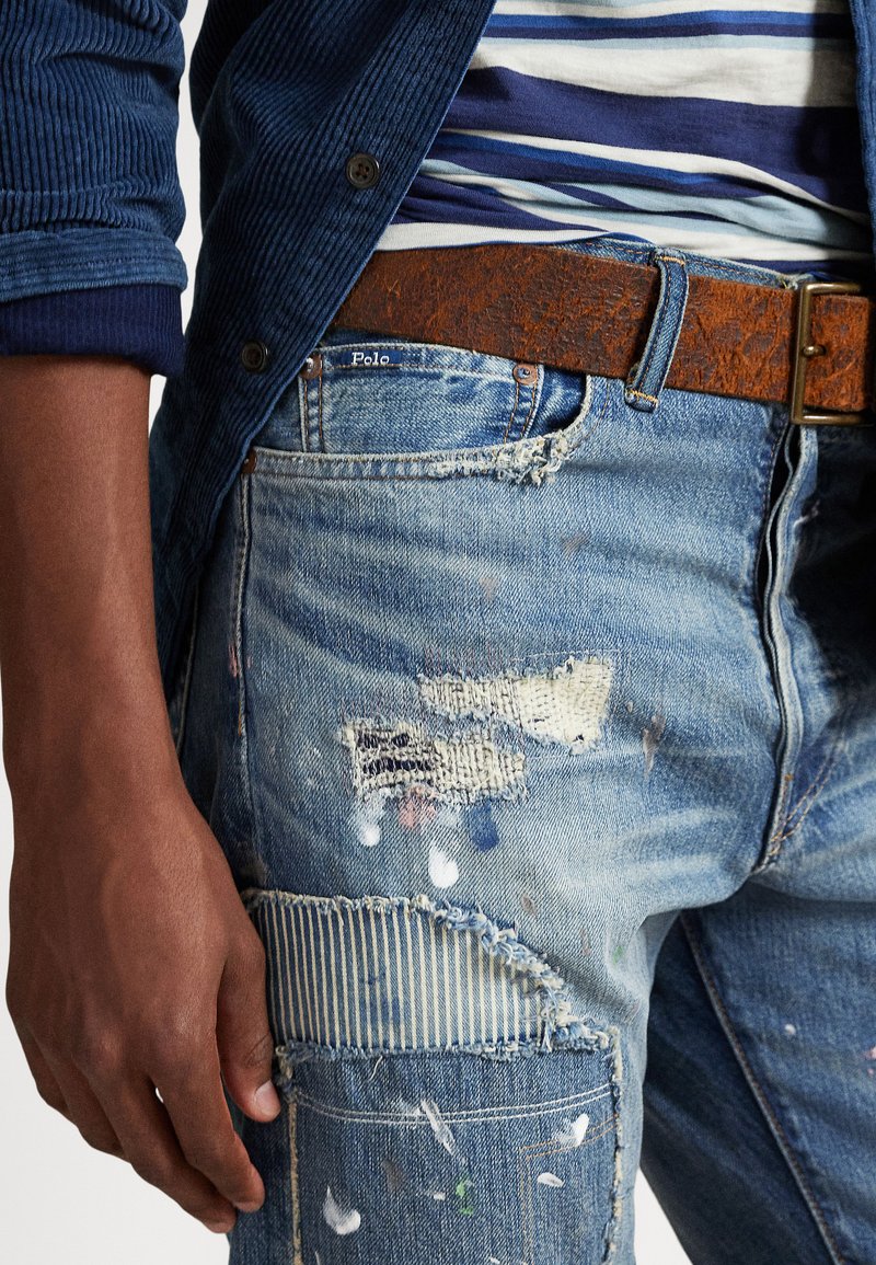 Primo piano di una persona che indossa jeans blu rattoppati e consumati con schizzi di vernice, una cintura di pelle marrone e una camicia a righe blu sotto una giacca blu.