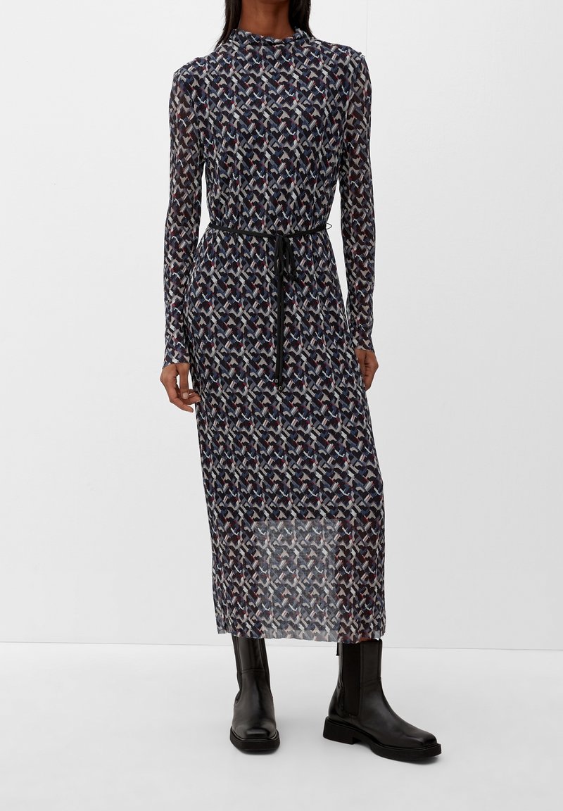 Femme portant une robe midi à manches longues à motifs avec une ceinture noire, associée à des bottines en cuir noir, sur un fond uni.