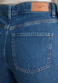Zbliżenie na tylną kieszeń niebieskich dżinsów z brązową metką marki Marc O’Polo Denim na pasku.