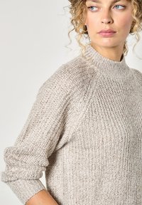 Helles beiges Strickpullover mit hohem Kragen, geripptem Material und aufgerollten Ärmeln, der einen weichen, lockeren Sitz und aufwendige Nähte zeigt.