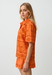 Oranje hemd met oogjes en golvende randen, bloemvormige uitsparingen, korte mouwen en een knoopsluiting. Draagt bijpassende shorts.