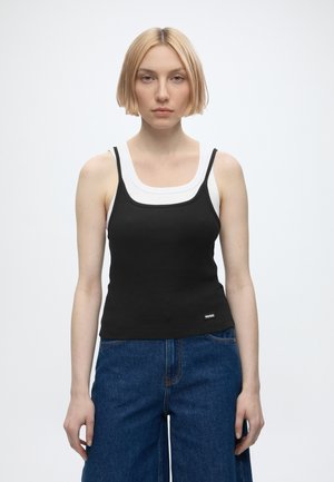 Jeune femme aux cheveux blonds courts, portant des débardeurs noirs et blancs superposés et un jean bleu, debout devant un fond clair uni.