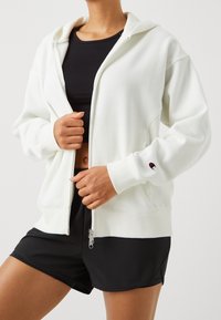 Sudadera blanca con cremallera y capucha, con puños acanalados, que presenta un pequeño logo en la manga, llevada sobre una camiseta negra y combinada con pantalones cortos negros.