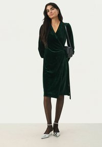 Grünes Samtwickelkleid mit langen Ärmeln, V-Ausschnitt und seitlichem Bindeband. Kombiniert mit transparenten Strumpfhosen und silbernen Spitzenschuhen.