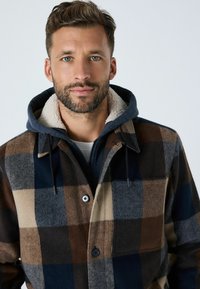 Ruudusline villane jakk, millel on pruun, tumesinine ja beež toon, nööpide sulgemisega ja fliisvoodriga kapuuts, et pakkuda lisasoojust.