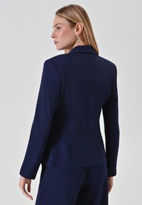 Marineblazer mit tailliertem Design, strukturierten Schultern und einem Peplum-Tailledetail. Glattes Material mit langen Ärmeln, ohne sichtbare Beschläge.
