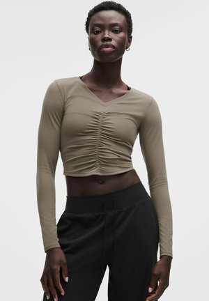 Vrouw met kort haar draagt een aansluitende, lange mouw, geplooide olijfgroene cropped top en hooggetailleerde zwarte broek tegen een effen achtergrond.