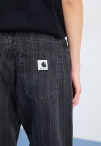 Pantaloni in denim a righe nere con una tasca posteriore che presenta un'etichetta bianca con il logo Carhartt, in mostra con una trama tessile e un design a taglio dritto.