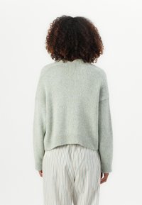 Persoon met krullend haar draagt een lichtgrijze trui en witte broek met dunne blauwe verticale strepen, staat met de rug naar de camera op een witte achtergrond.