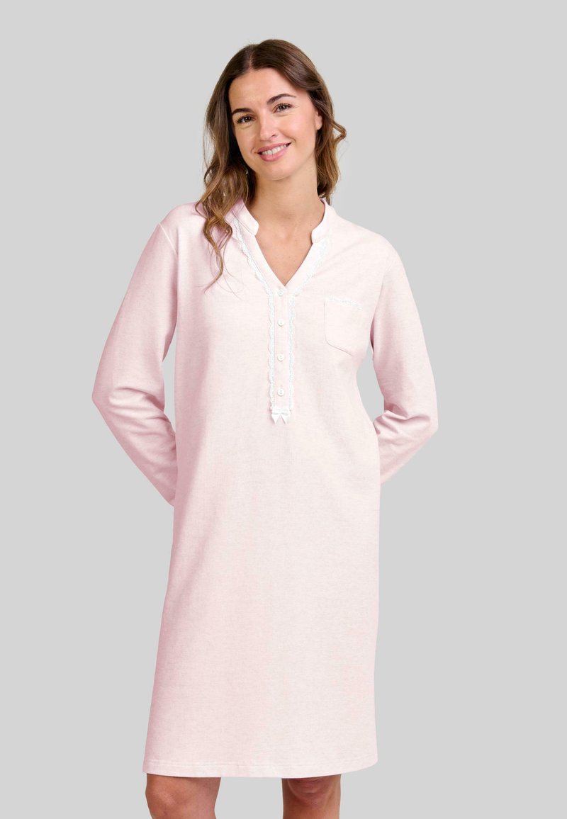 Mujer sonriendo, vistiendo un vestido de noche de manga larga en un tono rosado claro, con un borde de encaje abotonado y un pequeño bolsillo en el pecho, de pie frente a un fondo liso.