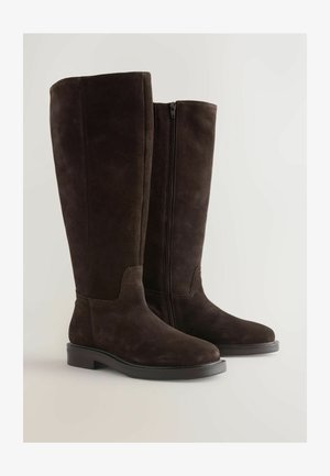 Brune ankelstøvler i ruskind med høj højde ved knæene har en glat overflade, rund tå, dekorativ syning og en lynlås på siden til lukning. Flad gummisål.