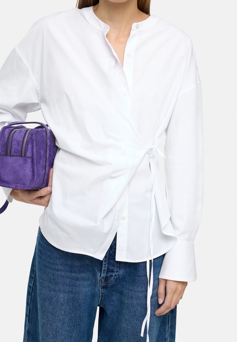 Biała koszula z długim rękawem i detalem w postaci bocznego wiązania, w połączeniu z małą fioletową torebką na ramię z zamszu. Całość dopełniają dżinsowe spodnie.