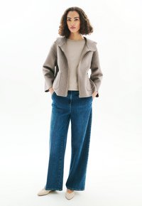 Veste gris clair en mélange de laine avec un large col et une fermeture éclair ; associée à un pantalon en denim bleu foncé ample et des chaussures légères beiges sans lacets.