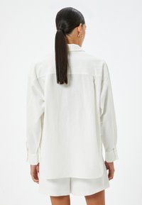 Camicia bianca a maniche lunghe con vestibilità rilassata, dotata di polsini abbottonati, yoke posteriore e orlo curvo. Tessuto morbido con texture minima.