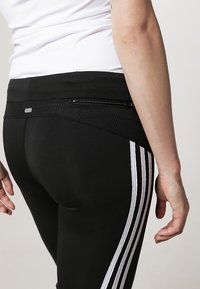 Černé sportovní legíny s hladkým povrchem, s bílými vertikálními pruhy po stranách a malou zipovou kapsou na zádech.