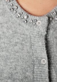 Lichtgrijs gebreid vest met kleine witte knopen en heldere kristallen versieringen langs de halslijn, waarbij de bovenborst en nek zichtbaar zijn.