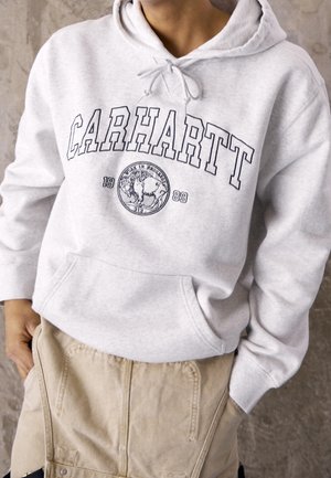 Persoon draagt lichtgrijze Carhartt hoodie met logo en afbeelding van een buffel boven zandkleurige broek, staand tegen een neutrale achtergrond.