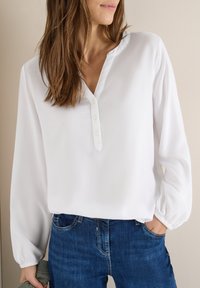 Blouse blanche en tissu lisse, avec une patte de boutonnage, des manches longues avec des poignets élastiques, portée avec un jean bleu foncé.
