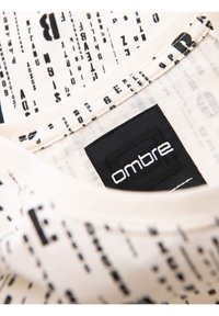 Tričko s límcem v krémové barvě s černým potiskem. Obsahuje černou značkovou etiketu s bílým nápisem "ombre".