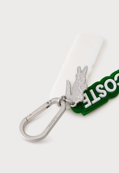 Сребърен карабинер ключодържател с гравирано "LACOSTE,", прикрепен към текстуриран сребрист крокодилов талисман и зелен гумен етикет "LACOSTE".