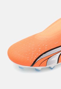 Jasnopomarańczowy but do piłki nożnej z teksturowaną cholewką, z czarnymi i białymi akcentami. Lekka konstrukcja i niebieskie plastikowe kołki na podeszwie.