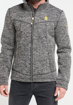 Mężczyzna noszący szarą dzianinową kurtkę zapinaną na zamek z żółtym logo na piersi i lewym rękawie, połączoną z ciemnoszarymi spodniami, pokazany od klatki piersiowej do bioder.