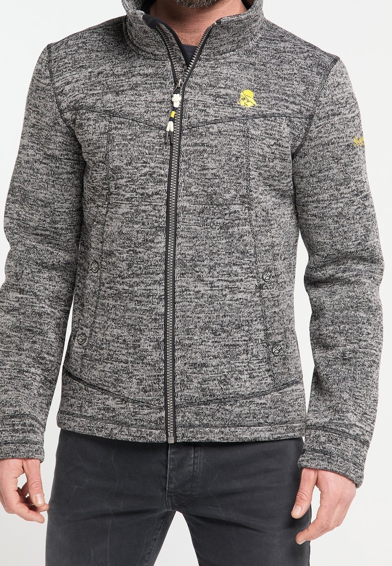 Homme portant une veste en tricot grise zippée avec un logo jaune sur la poitrine et la manche gauche, associée à un pantalon gris foncé, visible du buste aux hanches.