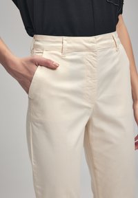 Cream-colored trousers in a smooth fabric, featuring side pockets and a mid-rise waist. A hand rests on the pocket, showcasing the fit.