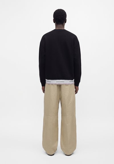 Homme portant un haut noir à manches longues et un pantalon beige ample, debout de dos sur un fond blanc uni.