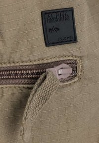 Beiger Stoff mit einer Gitterstruktur zeigt ein schwarzes Alpha Industries-Label. Ein beiger Reißverschlussgriff und braune Reißverschlusszähne sind sichtbar.