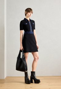 Robe noire à manches courtes avec col en denim et poches, fermeture éclair à l'avant et ceinture en denim. Le mannequin tient un sac noir plissé et porte des bottes à talons épais.